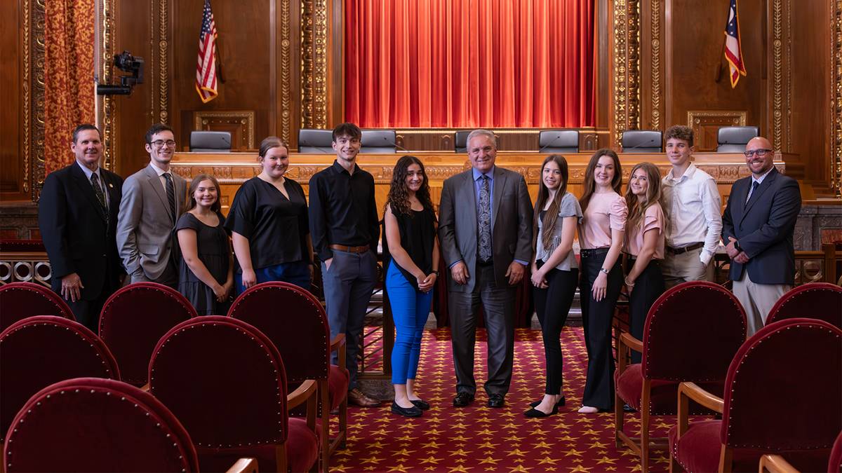 A group of students, teachers, and court staff stand in front of red curtains in the courtroom of the Supreme Court of Ohio.